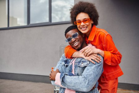 Smiling young african american woman in sunglasses and modern outfit embracing best friend and looking at camera while standing near building on urban street, stylish friends in city