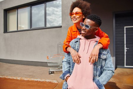 Smiling young african american woman in sunglasses and bright clothes embracing fashionable best friend in denim jacket while spending time on urban street, stylish friends in city concept