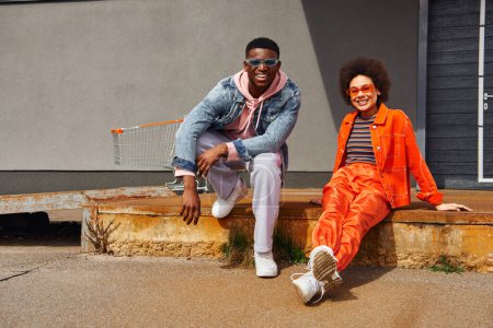 In voller Länge fröhliche und trendige junge afrikanisch-amerikanische beste Freunde in Sonnenbrille und modernem Outfit, die in die Kamera schauen, während sie auf rostigen Treppen in der Nähe von Gebäuden im Freien posieren, stilvolle Freunde in der Stadt