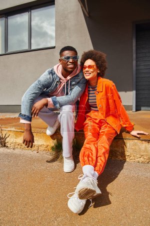 Lächelnde junge afrikanisch-amerikanische beste Freunde in Sonnenbrille und heller Kleidung sitzen auf rostigen Treppen in der Nähe von Gebäuden im Hintergrund und verbringen Zeit auf urbanen Straßen, stilvolle Freunde in der Stadt