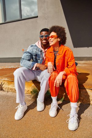 Lächelnder junger afrikanisch-amerikanischer Mann mit Sonnenbrille umarmt trendige beste Freundin in hellem Outfit, während er auf rostigen Treppen in der Nähe eines Gebäudes an der Stadtstraße sitzt, stilvolle Freunde in der Stadt