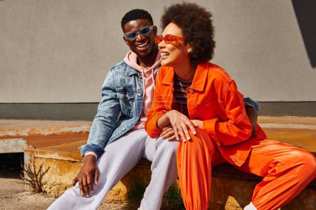 Fröhliche junge afrikanisch-amerikanische beste Freunde in trendigen Outfits und Sonnenbrille, die lachen und Spaß haben, während sie auf rostigen Treppen in der Nähe von Gebäuden an der Stadtstraße sitzen, trendige Freunde in urbanen Umgebungen