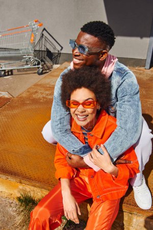 Pleased young african american man in sunglasses and trendy outfit embracing stylish best friend and sitting on rusty stairs near building on urban street , trendy friends in urban settings