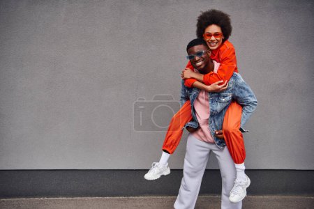 Lächelnde junge afrikanisch-amerikanische Frau in Sonnenbrille und hellem Outfit huckepack auf stylischem besten Freund und blickt in die Kamera in der Nähe eines Gebäudes an der Stadtstraße, trendige Freunde in urbaner Umgebung