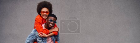 Foto de Positiva joven afroamericana mujer en gafas de sol y traje brillante piggybacking en mejor amigo y de pie cerca del edificio en la calle urbana, amigos de moda en entornos urbanos, pancarta - Imagen libre de derechos
