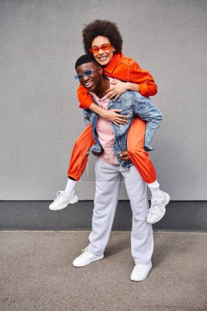 Full length of cheerful and stylish young african american woman piggybacking on best friend in sunglasses and denim jacket while standing near building outdoors, trendy friends in urban settings
