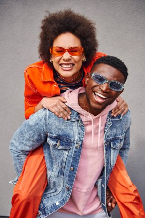Retrato de una joven afro-americana positiva y con estilo en gafas de sol apoyándose en el mejor amigo en chaqueta de mezclilla y mirando a la cámara cerca del edificio al aire libre, amigos de moda en entornos urbanos