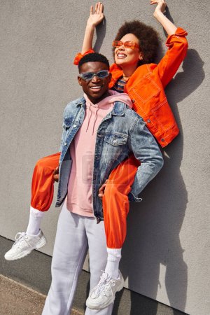 Positive and trendy young african american man in denim jacket holding best friend in sunglasses and bright outfit and standing near building on urban street, trendy friends in urban settings