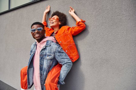 Feliz joven afroamericano hombre en gafas de sol y chaqueta de mezclilla sosteniendo mejor amigo en traje brillante y divertirse cerca de la pared en la calle urbana, amigos de moda en entornos urbanos