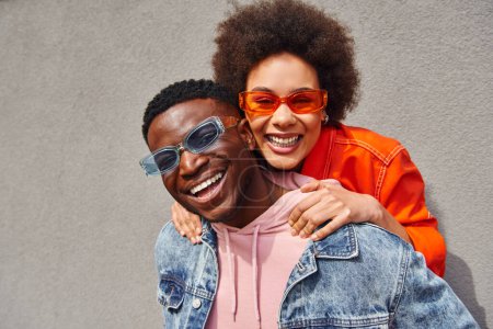 Porträt einer fröhlichen jungen afrikanisch-amerikanischen Frau mit natürlichen Haaren, die ihren besten Freund in stylischer Sonnenbrille und Jeansjacke umarmt, während sie in der Nähe von Gebäuden in die Kamera blickt, trendige Freunde in städtischen Umgebungen