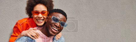 Foto de Mujer americana africana alegre en gafas de sol modernas y traje brillante abrazando mejor amigo y mirando a la cámara mientras está de pie cerca del edificio en la calle urbana, amigos con estética de moda, pancarta - Imagen libre de derechos