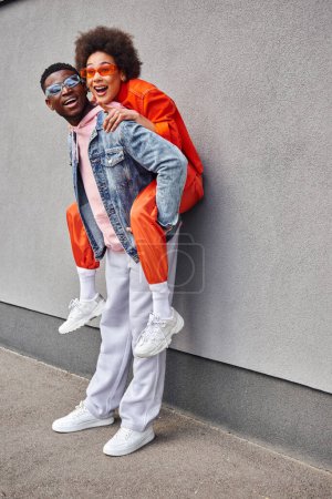 Excited african american woman in sunglasses and stylish outfit pointing with finger and piggybacking on best friend while standing near building on urban street, friends with trendy aesthetic
