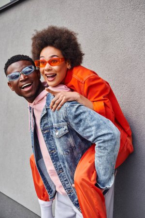 Cheerful young african american woman in stylish sunglasses pointing with finger while piggybacking on best friend and standing near building on urban street, friends with trendy aesthetic