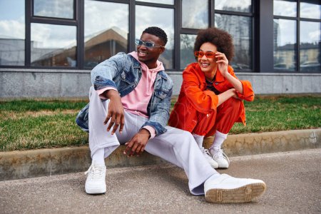 In voller Länge lächelnde und trendige junge afrikanisch-amerikanische beste Freunde mit Sonnenbrille, die wegschauen, während sie nebeneinander auf verschwommenen urbanen Straßen sitzen, Freunde mit trendiger Ästhetik