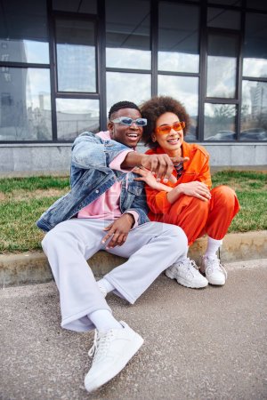 Téléchargez les photos : Jeune homme afro-américain souriant et branché avec des lunettes de soleil pointant du doigt tout en étant assis à la frontière à côté du meilleur ami en tenue élégante dans la rue urbaine, amis avec esthétique à la mode - en image libre de droit