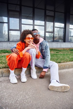 Full length of positive young african american man in sunglasses hugging best friend in bright outfit and sitting together on border on urban street, friends with trendy aesthetic