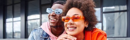 Mujer afroamericana joven y positiva con cabello natural que usa gafas de sol elegantes y mira hacia otro lado cerca de su mejor amigo mientras pasa tiempo en la calle urbana, amigos con estética de moda, pancarta 