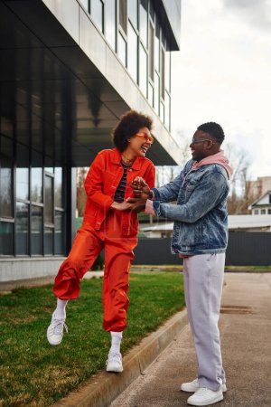 Positive young african american woman in sunglasses and bright outfit holding hands of stylish best friend and walking on border on urban street, stylish friends enjoying company concept