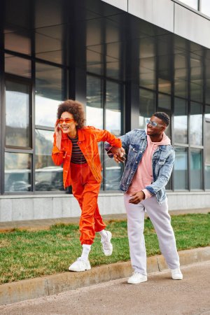 Full length of cheerful and modern african american woman in sunglasses and bright outfit walking on border and holding hand of best friend on urban street, stylish friends enjoying company