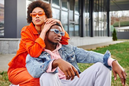 Moderne junge Afroamerikanerin in Sonnenbrille und hellem Outfit umarmt stilvolle beste Freundin, während sie posiert und Zeit auf urbaner Straße verbringt, stilvolle Freunde genießen Firmenkonzept