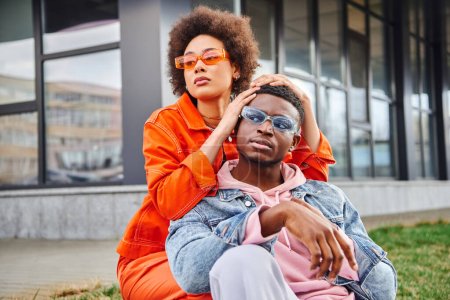 Confident and stylish young african american woman in sunglasses and bright outfit posing with best fried and looking away while spending time on urban street, stylish friends enjoying company