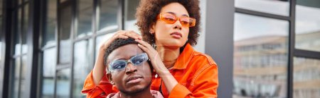 Confident and trendy young african american woman in sunglasses and bright outfit posing with best friend and looking away on blurred urban street, stylish friends enjoying company, banner 