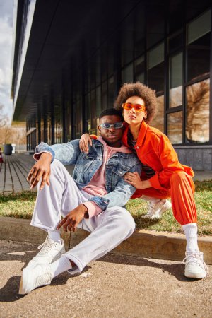 Full length of trendy and confident african american best friends in sunglasses looking at camera while sitting on border on blurred urban street at background, stylish friends enjoying company tote bag #664380980