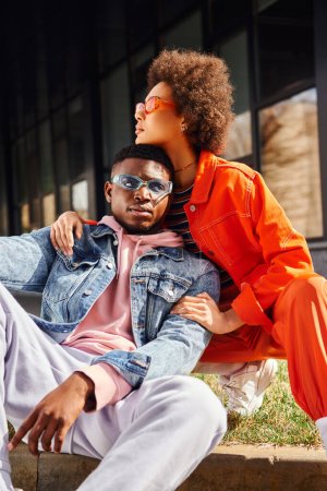 Modische junge afrikanisch-amerikanische Frau in Sonnenbrille und hellem Outfit umarmt beste Freundin sitzt an der Grenze auf verschwommenen städtischen Straße im Hintergrund, stilvolle Freunde genießen Gesellschaft