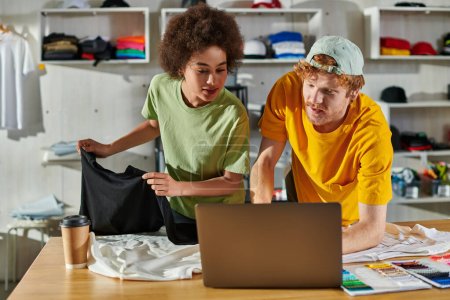 Artesanos jóvenes y multiétnicos mirando a la computadora portátil mientras trabajan junto con la ropa cerca del café para ir y muestras de color en la mesa en el estudio de impresión, concepto de éxito de la pequeña empresa