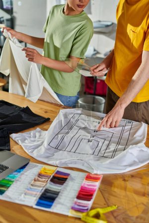 Ausgeschnittene Ansicht eines Handwerkers, der Druckmuster hält und auf T-Shirt zeigt, während er mit einem afrikanisch-amerikanischen Kollegen in der Nähe von Laptop und Farbmustern im Druckstudio arbeitet, Konzept für den Erfolg kleiner Unternehmen