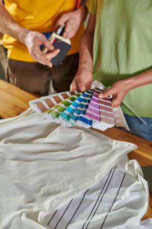 Cropped view of young african american craftswoman holding color swatches near colleague pointing with finger and standing near clothes in print studio, small business success concept