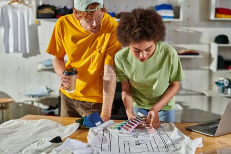Joven diseñador afroamericano sosteniendo muestras de color cerca de la ropa mientras trabaja con un colega sosteniendo café para ir y portátil en la mesa en el estudio de impresión, próspero concepto de pequeña empresa