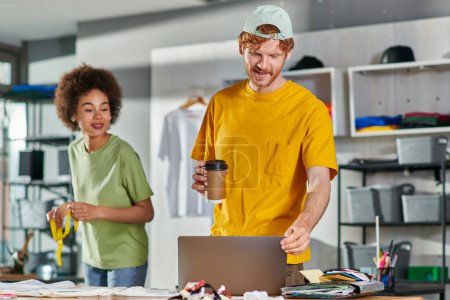 Smiling young designer holding coffee to go and using laptop while working near african american colleague with tape measure in blurred print studio, start-up innovation concept