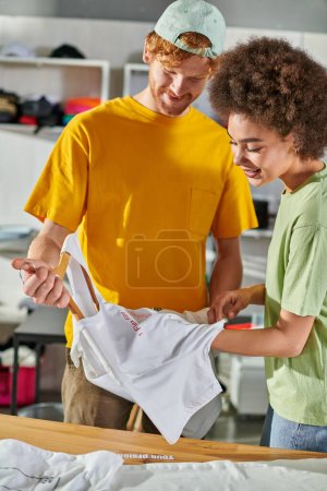 Smiling young african american designer holding t-shirt with lettering on hanger and working with colleague in blurred print studio at background, start-up innovation concept