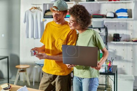 Smiling young craftsman holding printing layer while working with african american colleague holding laptop in blurred print studio at background, self-employment opportunity concept