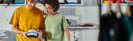 Photo for Cheerful young multiethnic craftspeople holding snapback with printing layer while working in blurred print studio at background, sustainable business model concept, banner - Royalty Free Image