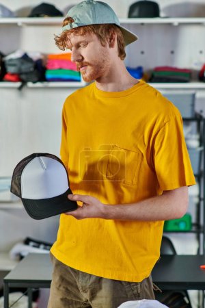 Photo for Young redhead craftsman holding snapback while working on project and standing in blurred print studio at background, small business resilience concept - Royalty Free Image