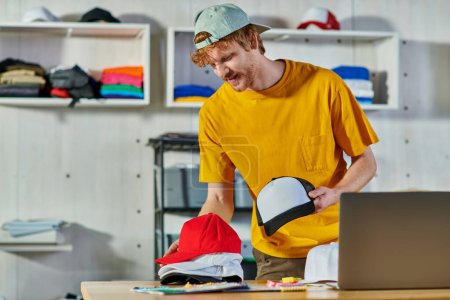 Cheerful young redhead craftsman holding snapbacks while working near cloth samples and blurred laptop on wooden table in print studio, small business resilience concept
