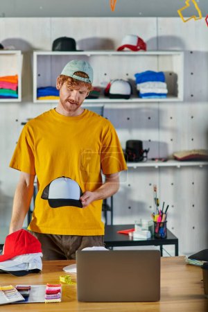 Young redhead craftsman holding snapback while working on project near laptop and cloth samples on table and standing in blurred print studio, small business resilience concept