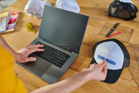 Cropped view of designer using laptop with blank screen and holding printing layer near snapback on blurred table in print studio, hands-on entrepreneurship concept 