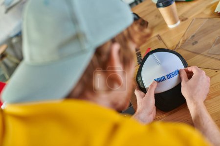 Photo for Blurred young designer holding printing layer near snapback while working near blurred coffee to go on table in print studio, hands-on entrepreneurship concept - Royalty Free Image