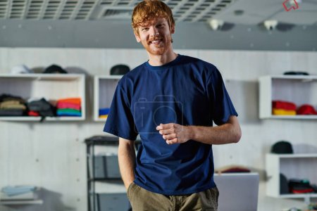 Portrait of positive young redhead craftsman in casual clothes looking at camera while posing and standing in blurred print studio, self-made success concept 