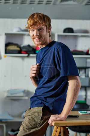 Portrait of young redhead craftsman in casual clothes looking at camera while standing near blurred smartphone on table in print studio, self-made success concept 