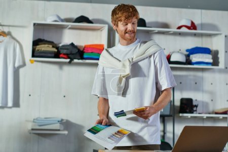 Smiling young redhead craftsman holding cloth samples while looking at laptop and working on project in print studio at background, self-made success concept 