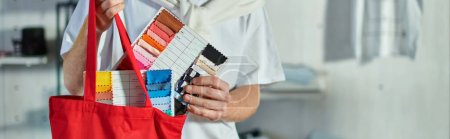 Cropped view of young craftsman in casual clothes putting cloth samples in shoulder bag in blurred print studio at background, self-made success concept, banner 