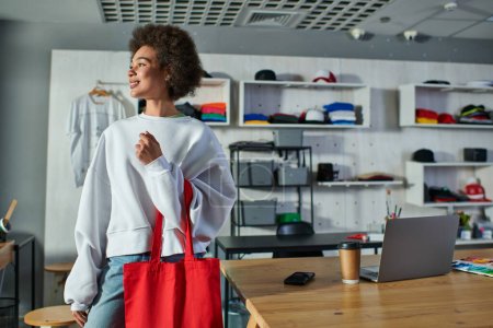 Smiling young african american craftswoman holding shoulder bag and looking away while standing near devices and coffee to go in print studio, enthusiastic business owner working in workshop