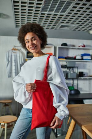 Cheerful young african american craftswoman holding shoulder bag and looking at camera while standing in blurred print studio, enthusiastic business owner working in workshop