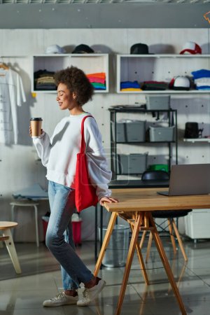 Téléchargez les photos : Vue latérale d'une jeune artisan afro-américaine positive avec sac à bandoulière tenant du café à emporter et debout près d'un ordinateur portable sur la table dans un studio d'impression, propriétaire d'entreprise enthousiaste travaillant dans un atelier - en image libre de droit