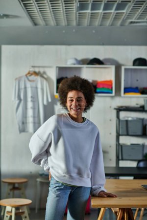 Fröhliche und selbstbewusste afrikanisch-amerikanische Handwerkerin in Sweatshirt und Jeans, die in die Kamera blickt, während sie am Arbeitstisch im Druckstudio steht, begeisterte Geschäftsinhaberin, die in der Werkstatt arbeitet