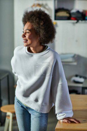 Portrait of cheerful young african american craftswoman in sweatshirt and jeans looking away while standing near working table in blurred print studio, enthusiastic business owner working in workshop
